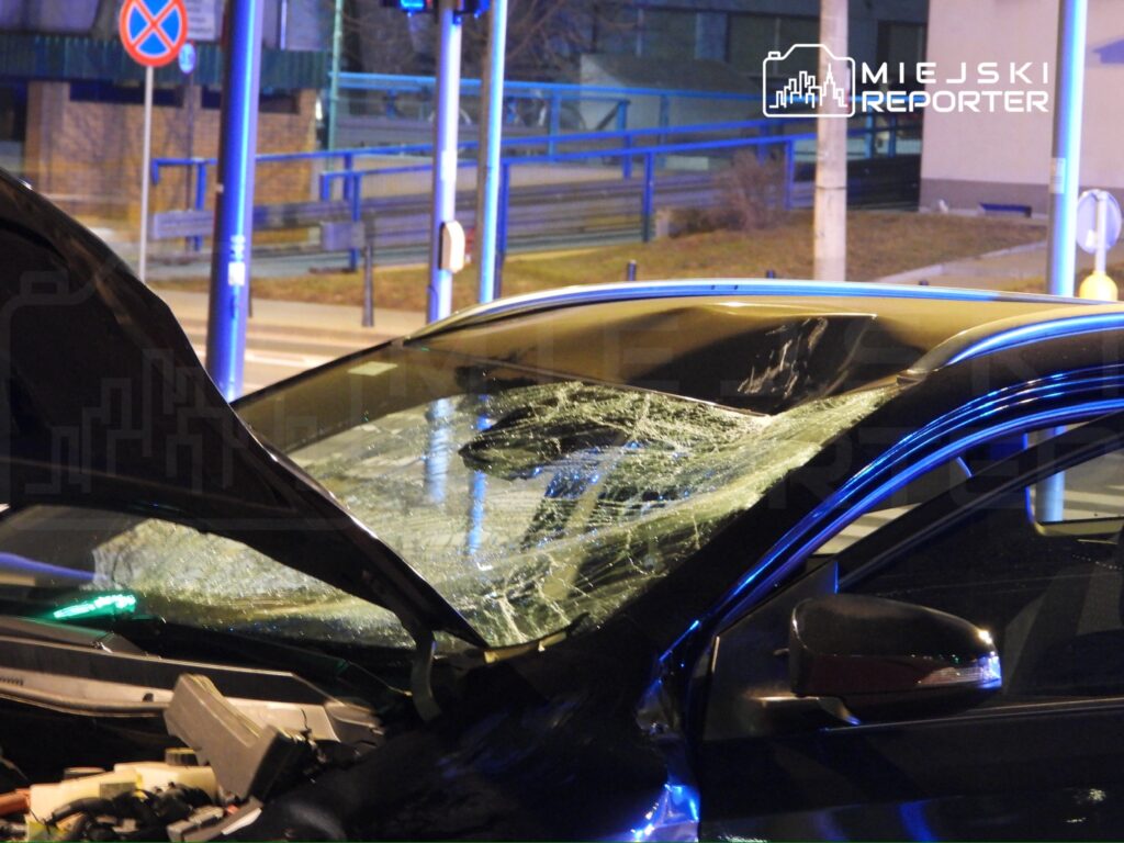 Uszkodzone auto z rozbitym przednią szybą stoi na ulicy w nocy, w tle widoczne są znaki drogowe i oświetlenie.
