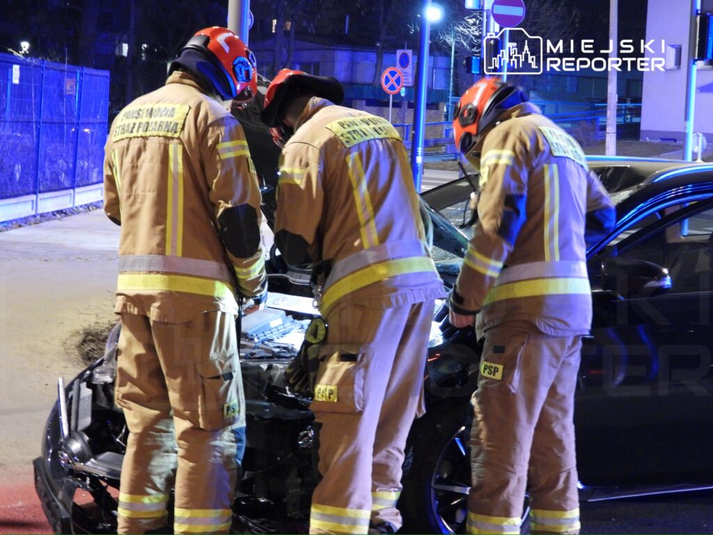 Trzech strażaków w mundurach ratunkowych pracuje przy uszkodzonym samochodzie w nocnej scenerii miejskiej.