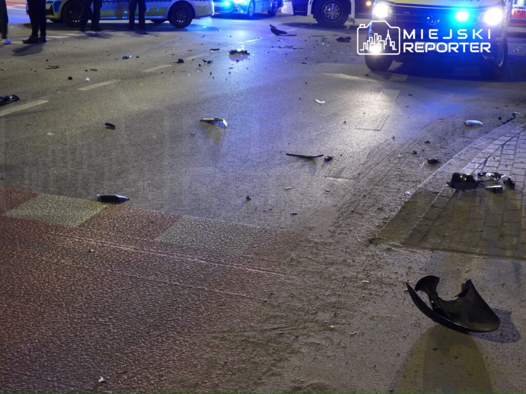 Na jezdni leżą fragmenty pojazdów oraz rozbite szkło, w tle widoczne policyjne radiowozy z włączonymi sygnałami świetlnymi.