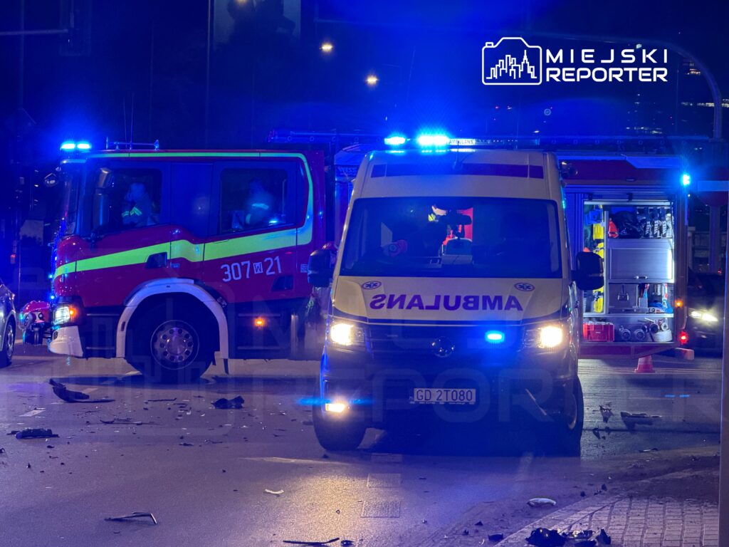 Czerwony wóz strażacki i ambulans z włączonymi sygnałami świetlnymi na oświetlonej ulicy, wokół rozbite fragmenty.