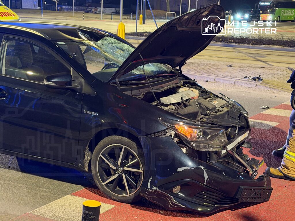 Uszkodzony czarny samochód osobowy z otwartą maską stoi na skrzyżowaniu, obok niego interweniujący strażacy.