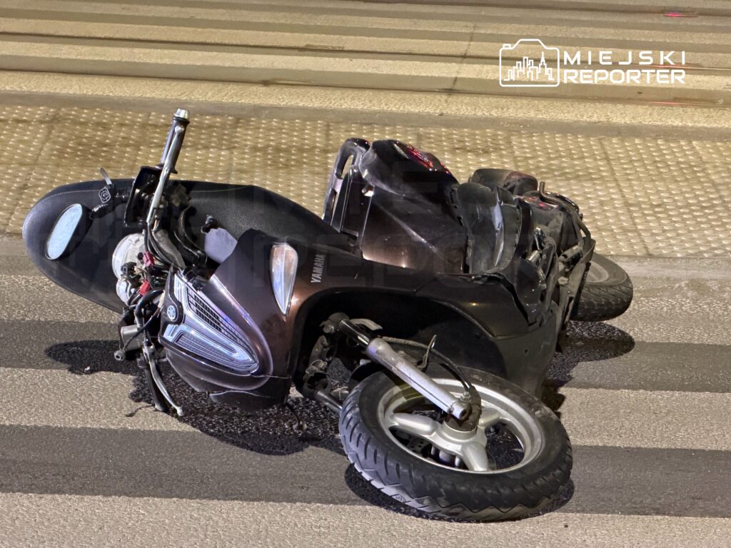 Uszkodzony skuter leży na jezdni, obok pasów dla pieszych, w nocy.