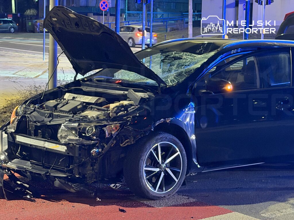 Czarny samochód osobowy z uszkodzoną maską i wgniecioną karoserią stojący na ulicy, obok latarni i znaków drogowych.