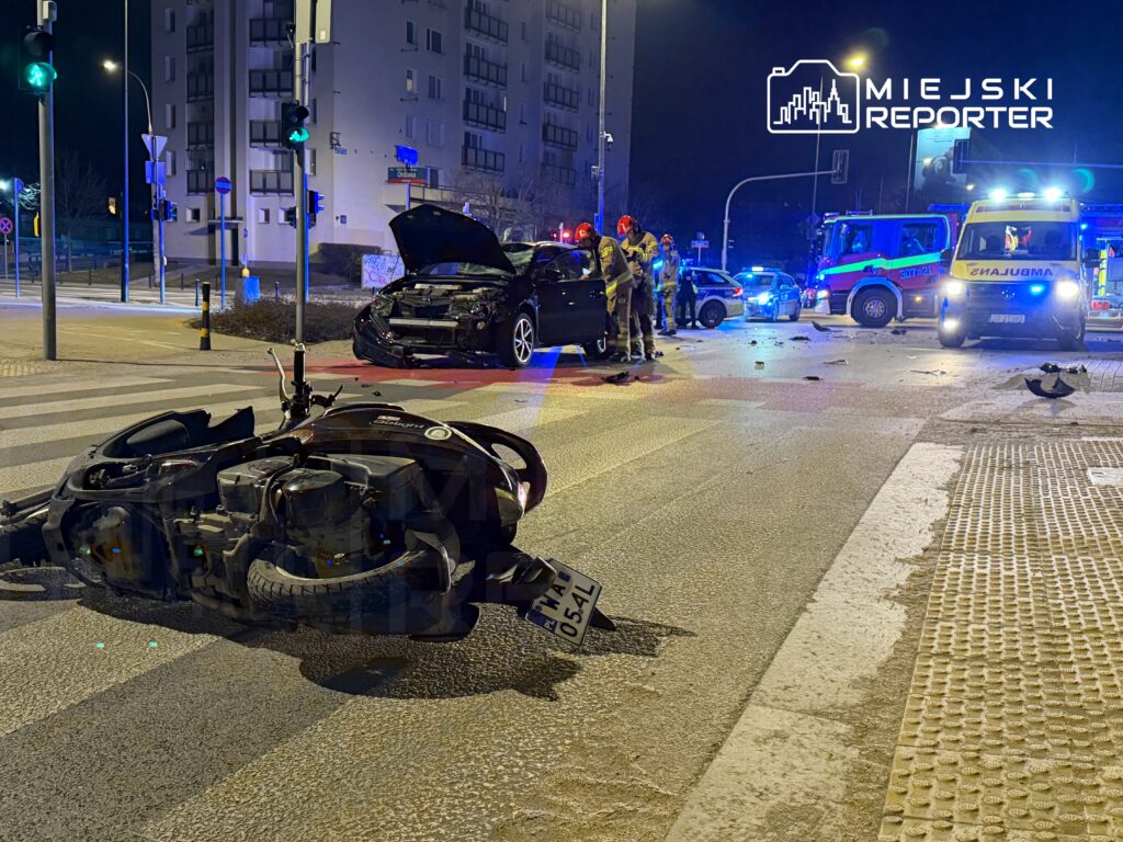 Na skrzyżowaniu leży przewrócony motocykl, obok uszkodzony samochód, a w tle widać interwencję służb ratunkowych.