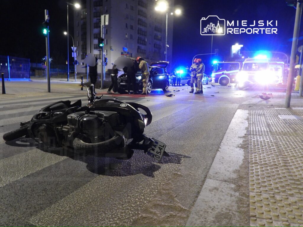 Motocykl leży na jezdni po wypadku, a w tle widać ratowników medycznych oraz policyjne samochody na oświetlonym skrzyżowaniu.