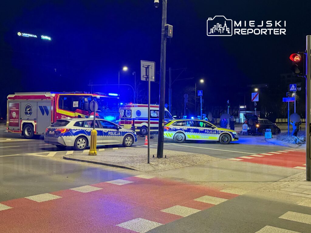 Na skrzyżowaniu stoją pojazdy służb ratunkowych, w tym wóz strażacki i radiowozy policyjne, w nocy z niebieskimi światłami.