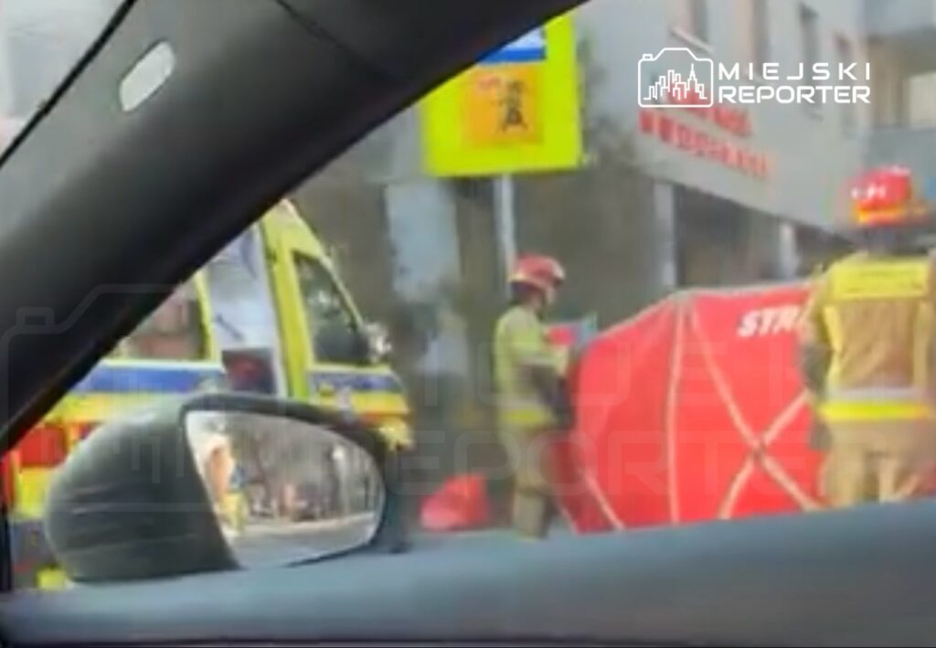 Strażacy w kaskach pracują przy czerwonym namiocie, obok karetki na ulicy w mieście.