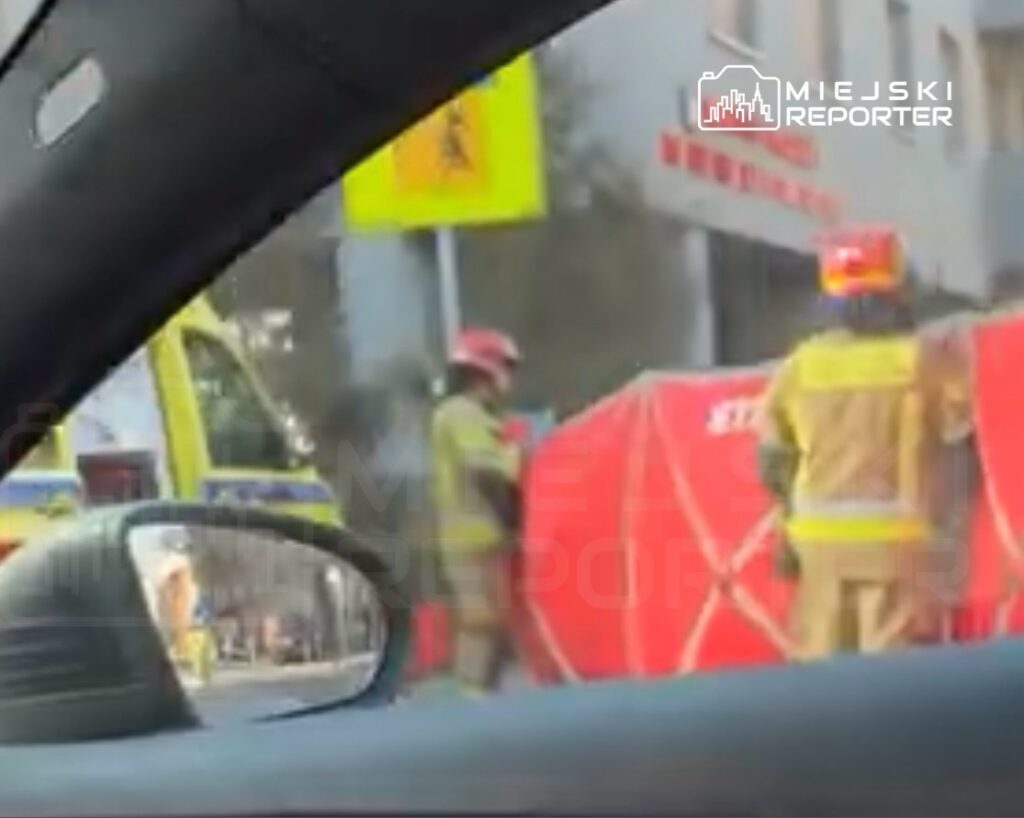 Strażacy w kaskach i mundurach zabezpieczają miejsce zdarzenia przy czerwonym namiocie. W tle widoczna karetka.