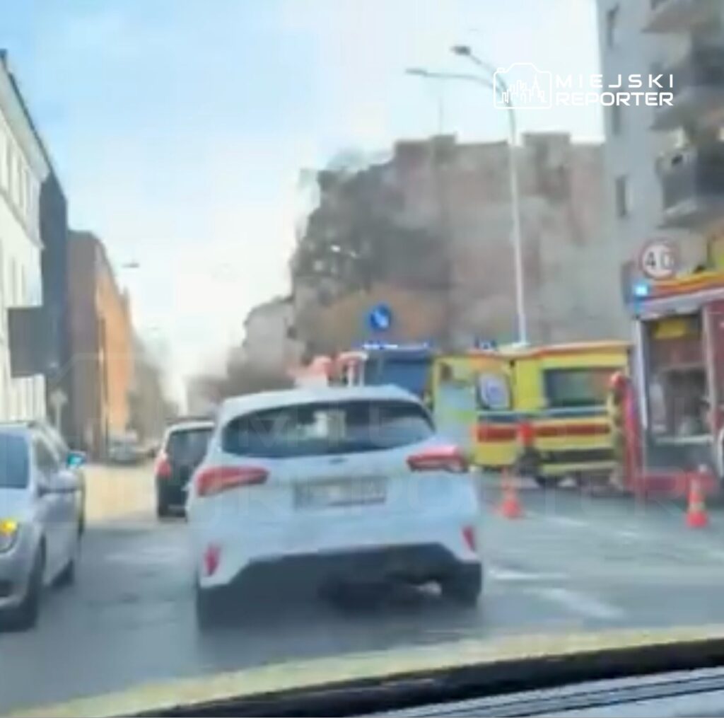 Kierowca w białym samochodzie przejeżdża obok pojazdów ratunkowych na zablokowanej ulicy.