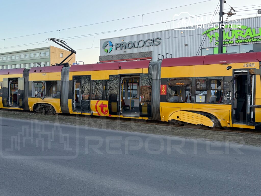 Uszkodzony tramwaj z otwartymi drzwiami stoi na torach obok nowoczesnego budynku z zielonym napisem.