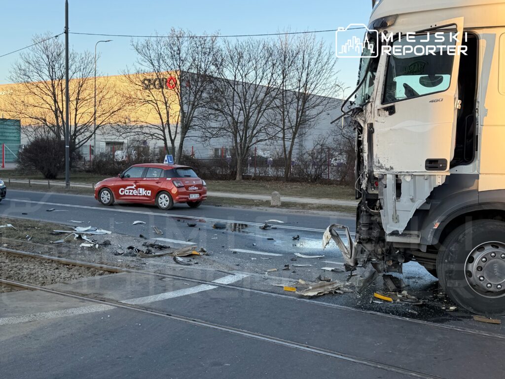 Wielki ciężarówka z uszkodzoną kabiną stoi na ulicy, obok leżą fragmenty rozbitego pojazdu oraz samochód szkoleniowy.
