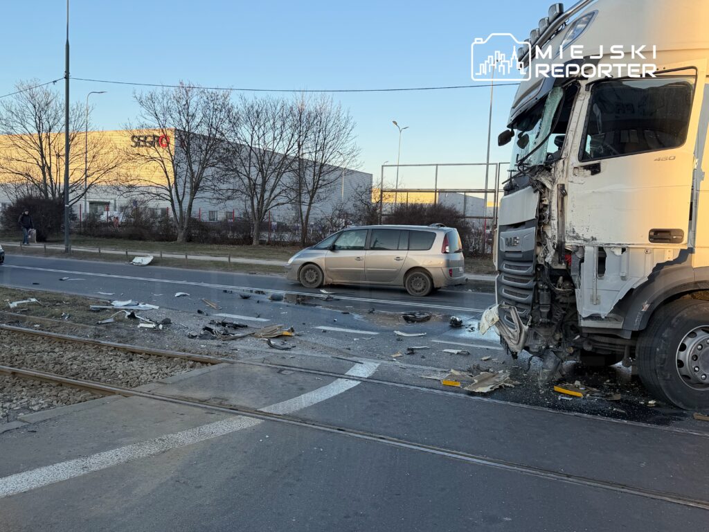 Zniszczona ciężarówka stojąca na drodze, obok leżące fragmenty wraku, w tle budynek przemysłowy.