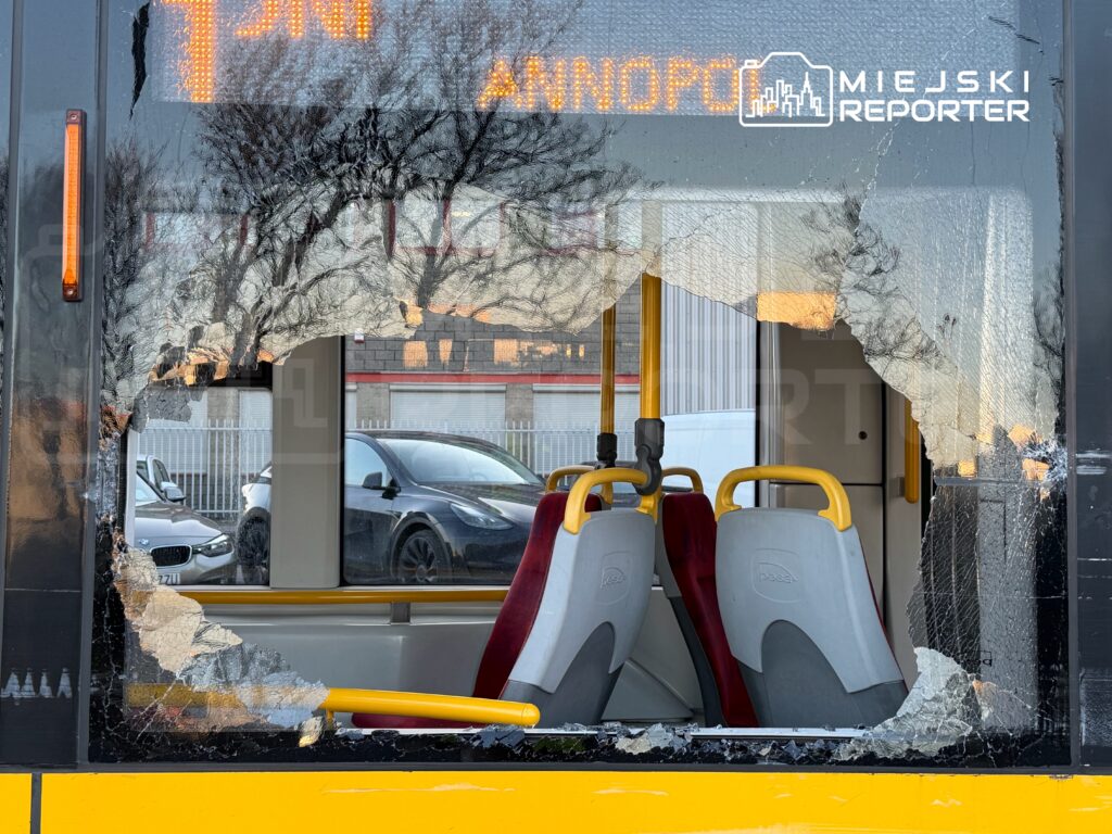 Zniszczona szyba autobusu z dużym otworem, w tle widać samochody zaparkowane przy ulicy.