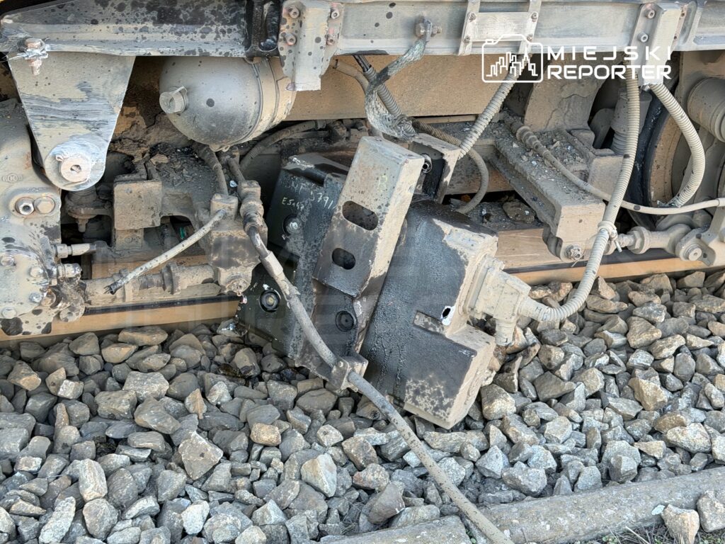 Uszkodzona część mechanizmu kolejowego leżąca na torach, otoczona kamieniami. Widoczne przewody i elementy metalowe.