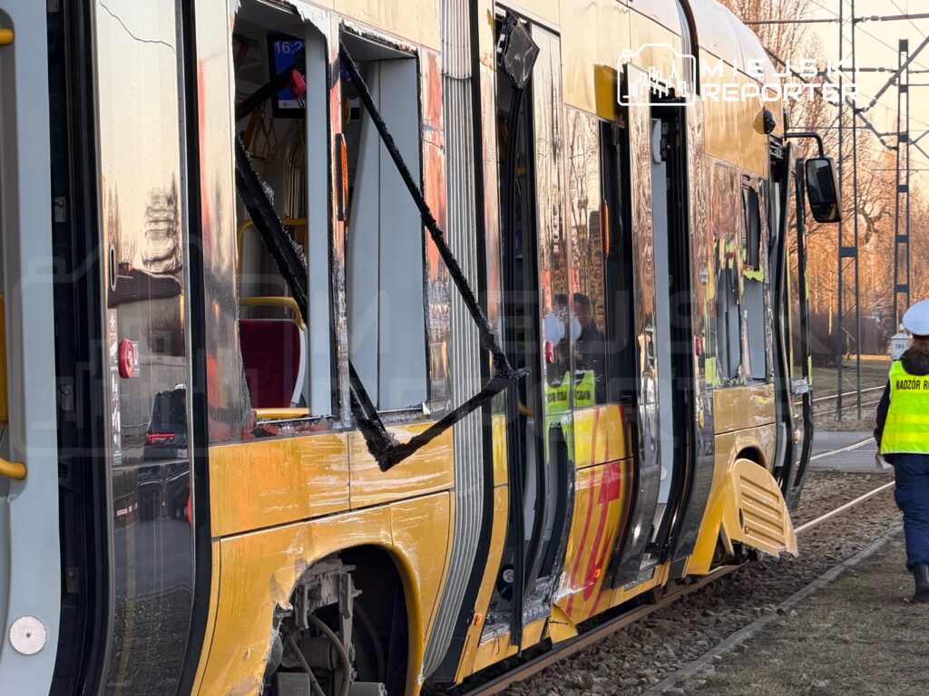 Uszkodzony tramwaj z wybitymi oknami stoi na torowisku, obok pracownicy w kamizelkach odblaskowych.