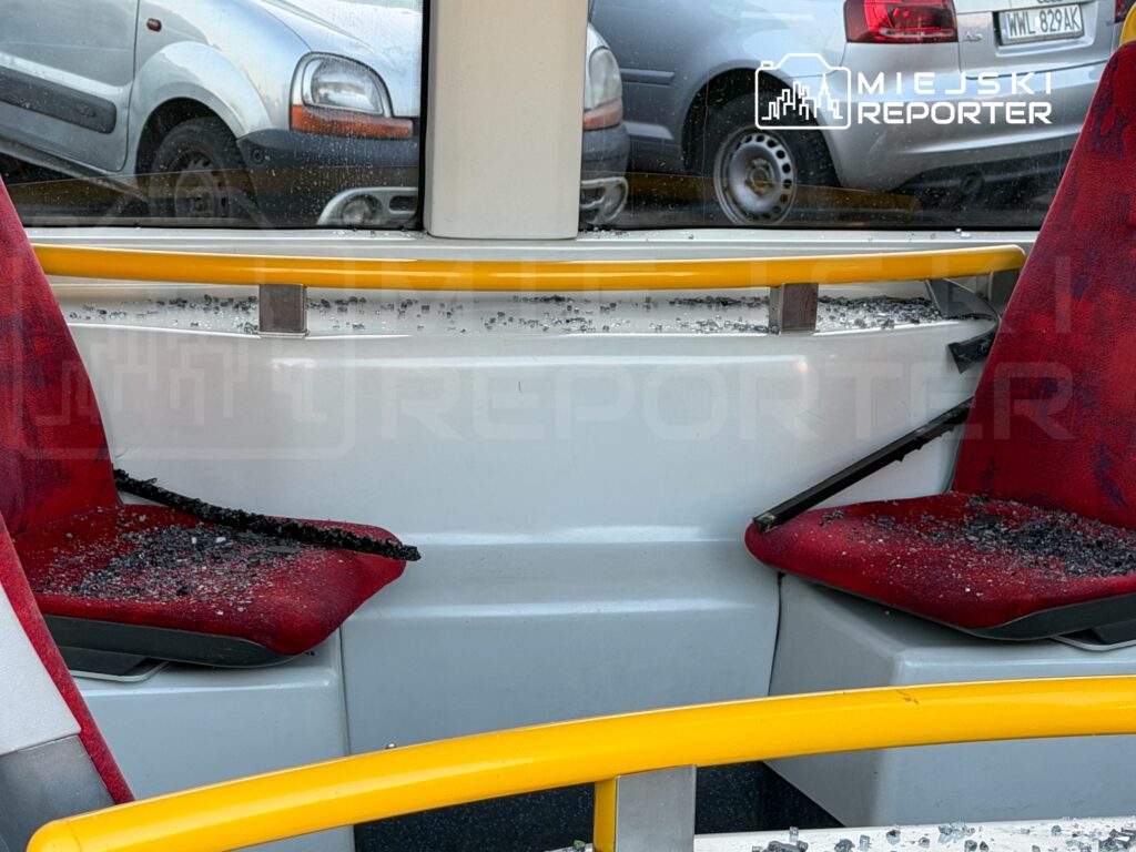 Zniszczone czerwone siedzenia w pojeździe komunikacji miejskiej, pokryte odłamkami szkła i zniszczonymi elementami wnętrza.