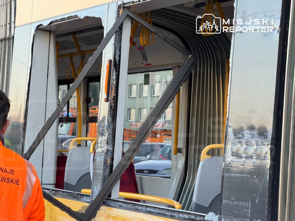 Uszkodzona ściana tramwaju z wyłamanym oknem, obok pracownik w pomarańczowej kamizelce. W tle widoczna ulica.