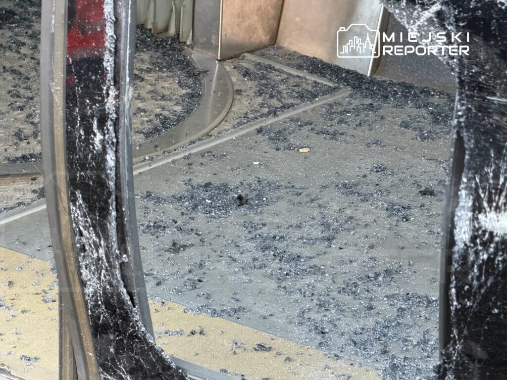 Szkło rozbite na podłodze w pomieszczeniu, z widocznymi kawałkami szkła i zarysowaniami na ścianie.