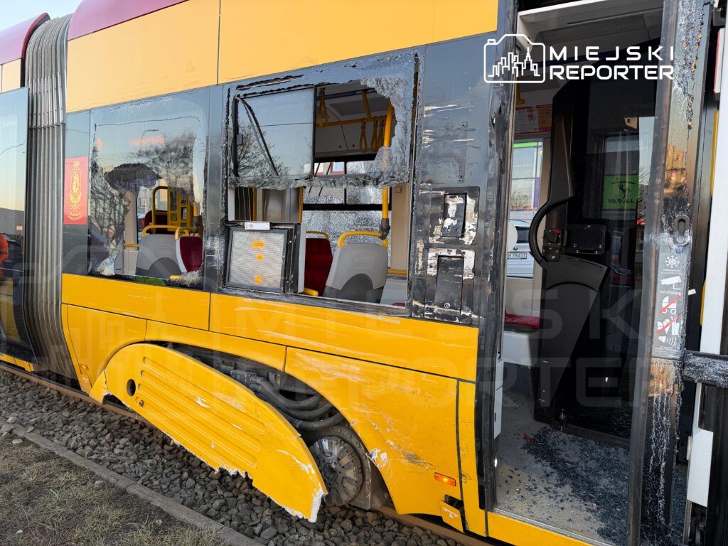 Uszkodzony tramwaj z wybitym oknem i zarysowaniami na karoserii stoi na torach w miejskim otoczeniu.