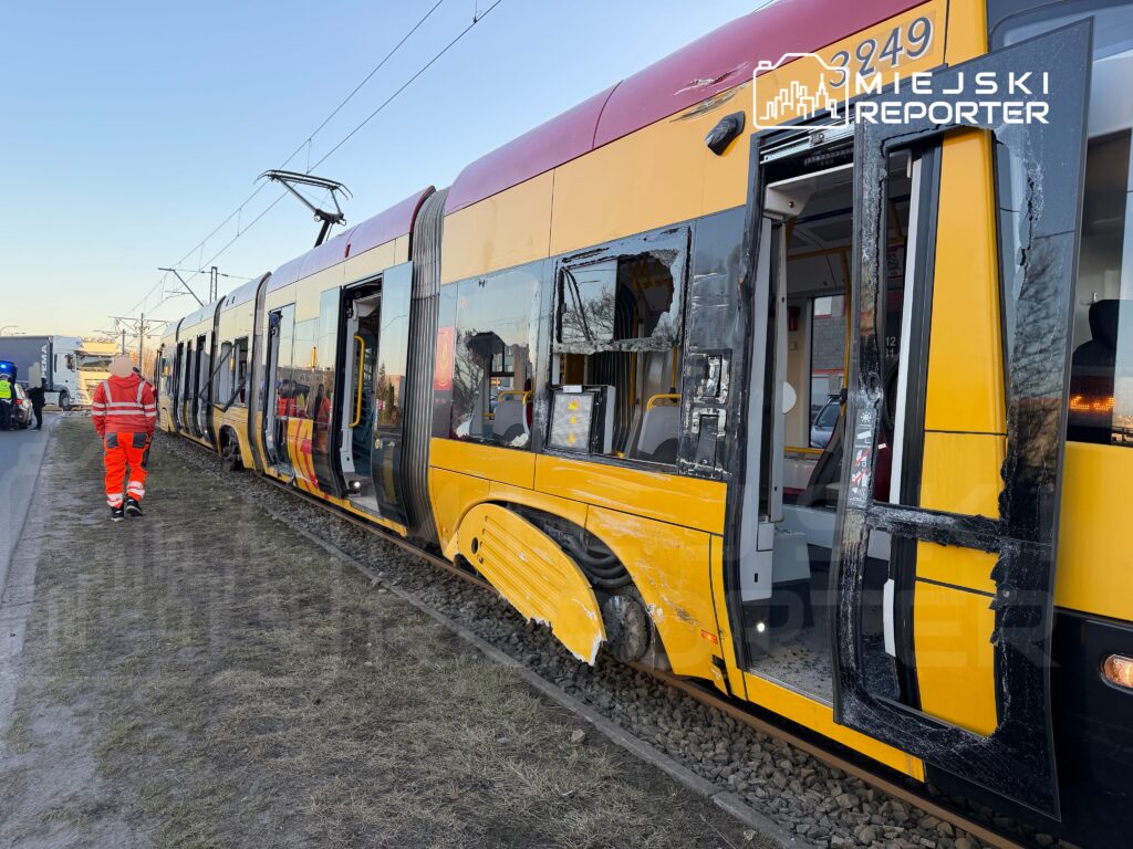 Zniszczony tramwaj stoi na torowisku, a pracownik w odzieży roboczej sprawdza uszkodzenia w pobliżu otwartych drzwi.
