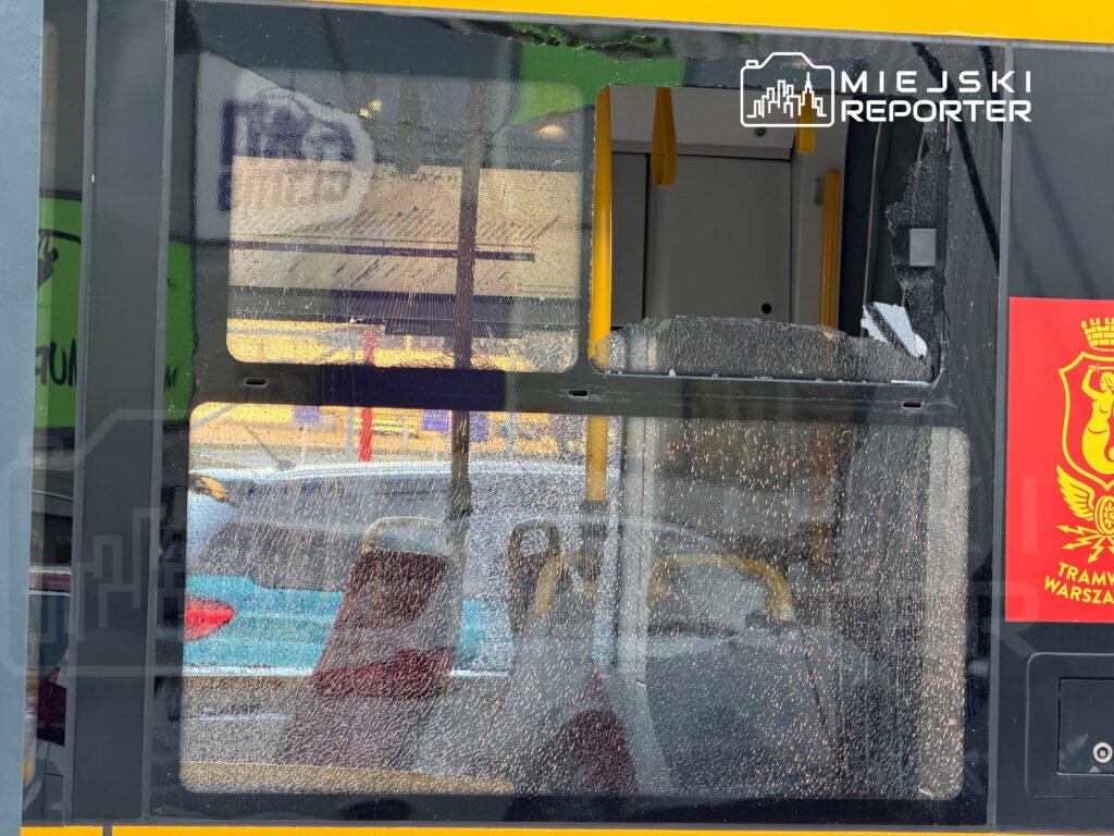 Zniszczona szyba w drzwiach tramwaju z widocznymi śladami uszkodzenia, w tle widać samochody na ulicy.