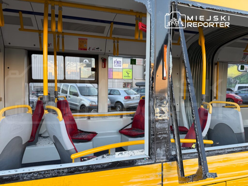 Wnętrze autobusu miejskiego z uszkodzonymi oknami i porozrzucanymi kawałkami szkła, czerwone siedzenia w tle.