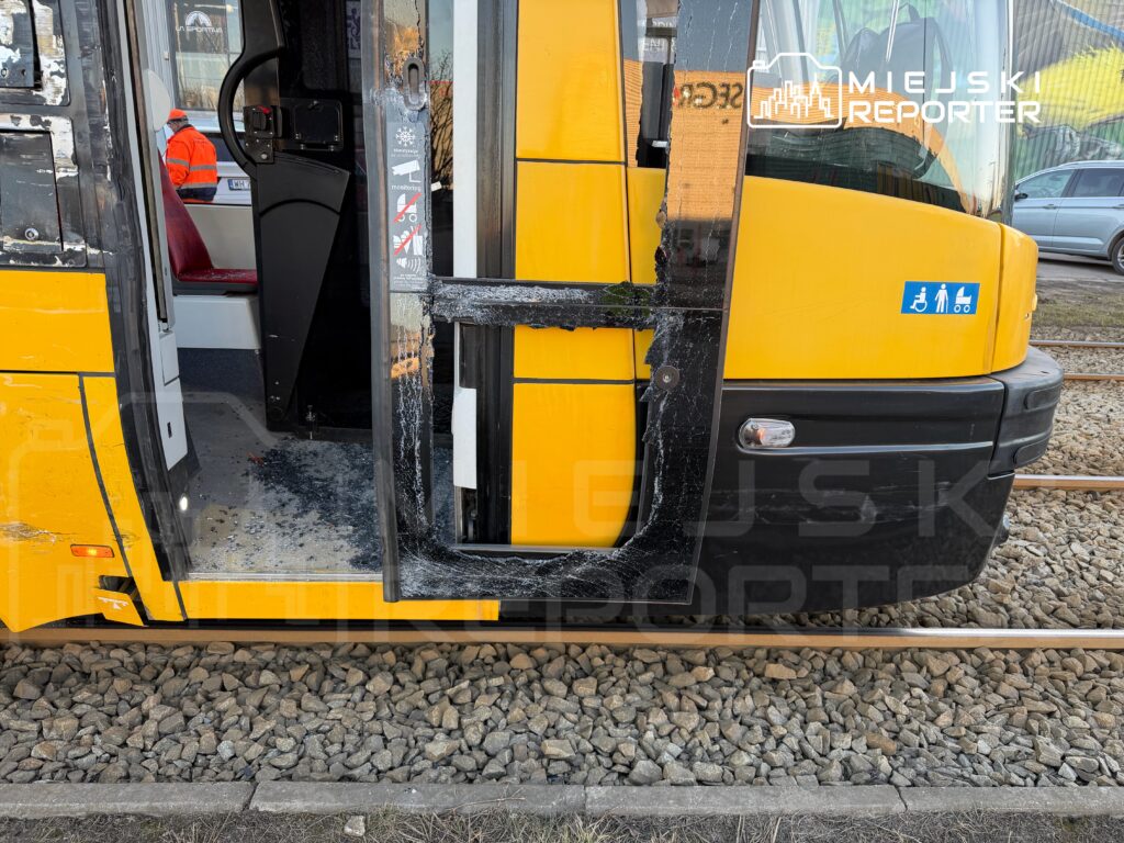 Otwarte drzwi żółtego tramwaju z uszkodzoną ramą, na tle torów i kamieni. Widać fragment wnętrza pojazdu.