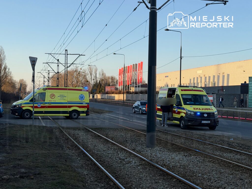 Ambulans z włączonymi sygnałami świetlnymi zatrzymany przy torach tramwajowych, obok przechodnia rozmawiającego z ratownikami.