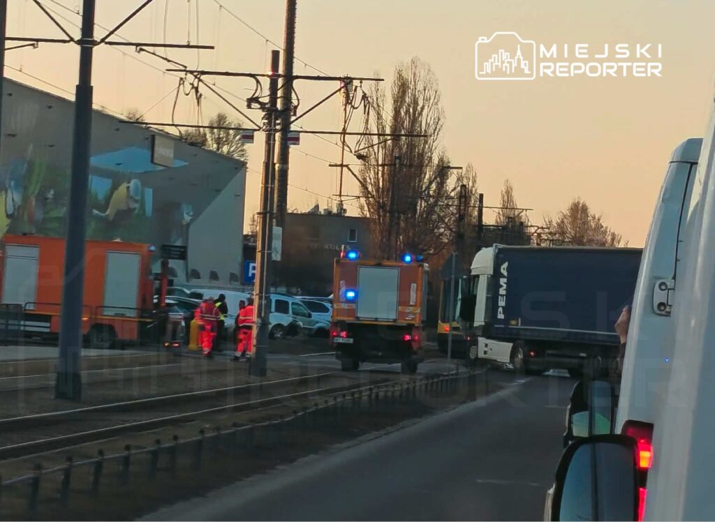 Grupa ratowników w pomarańczowych strojach pracuje obok wozu strażackiego przy torach tramwajowych o zachodzie słońca.