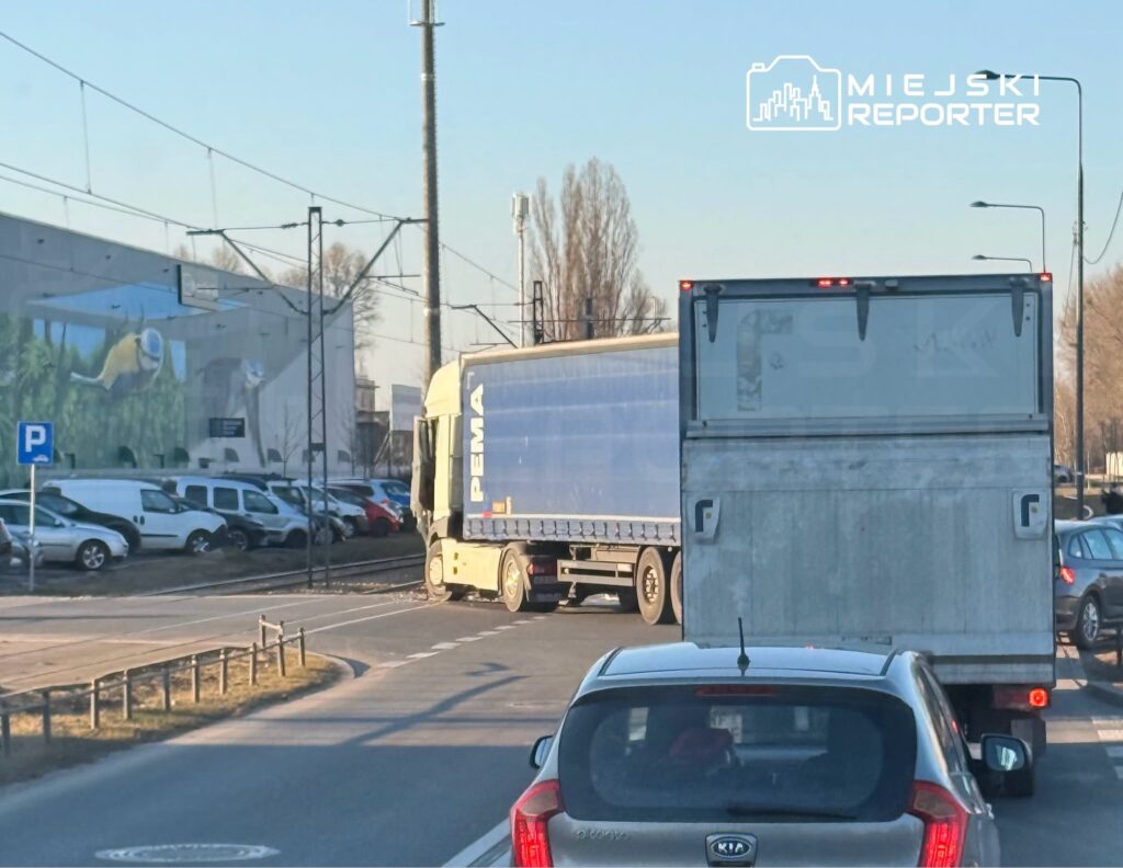 Tir skręcający w prawo na rondzie, w tle nowoczesny budynek z muralem oraz zaparkowane samochody.