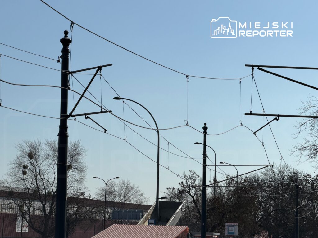 Linie tramwajowe zawieszone na słupach w tle błękitnego nieba, z widocznymi drzewami w okolicy.
