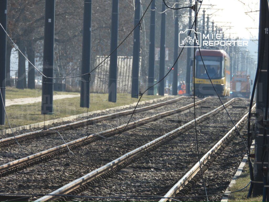 Tramwaj zbliżający się do stacji na torach, otoczony słupami trakcyjnymi i drzewami w tle.