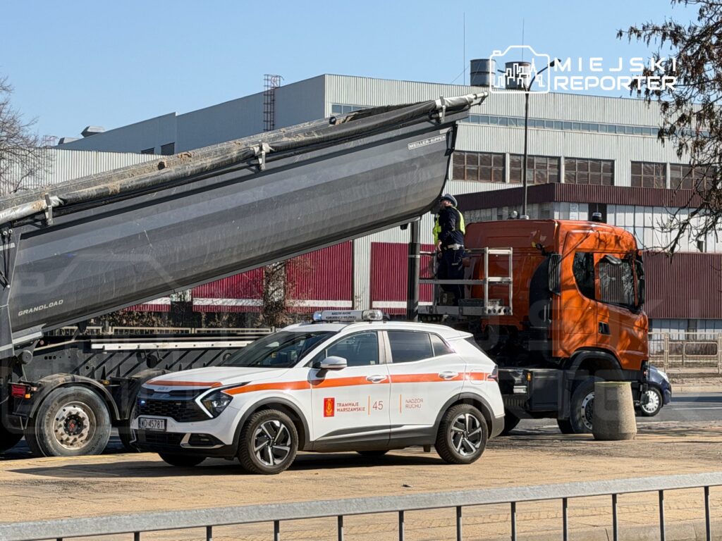 Pracownik w kamizelce odblaskowej kontroluje załadunek materiałów do wywrotki na placu budowy obok samochodu służb miejskich.