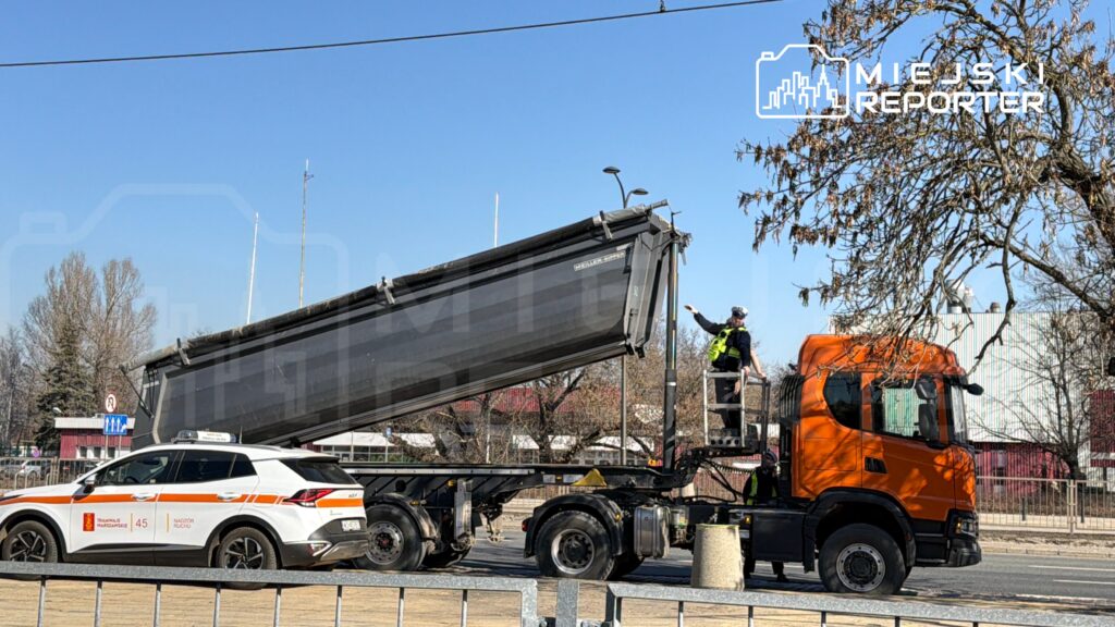 Mężczyzna w kamizelce odblaskowej kieruje podnoszeniem naczepy ciężarówki na ulicy, obok samochodu patrolowego.