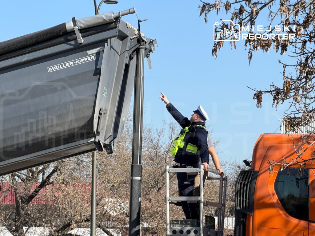 Policjant w mundurze wskazuje coś z wysokości platformy obok podnoszonej przyczepy.