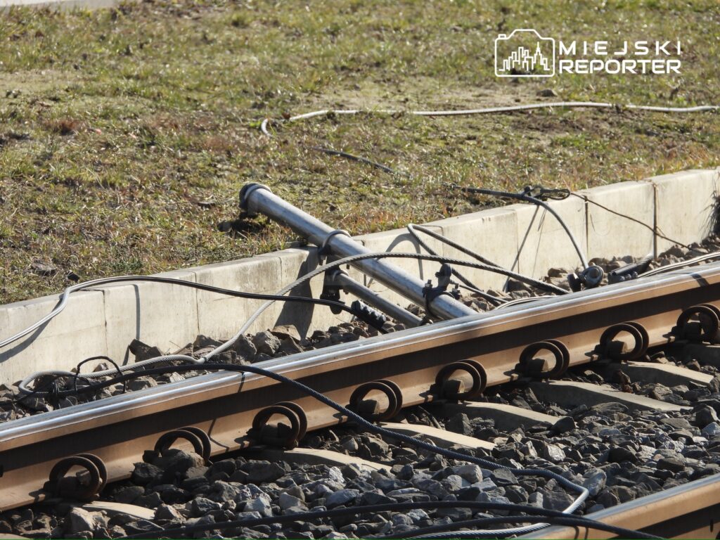 Uszkodzone tory kolejowe z wystającymi elementami i porozrzucanymi kablami na trawie obok torowiska.