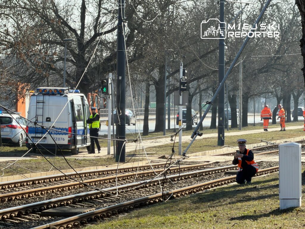 Policjant w kamizelce odblaskowej obserwuje uszkodzone linie tramwajowe obok zaparkowanego radiowozu.