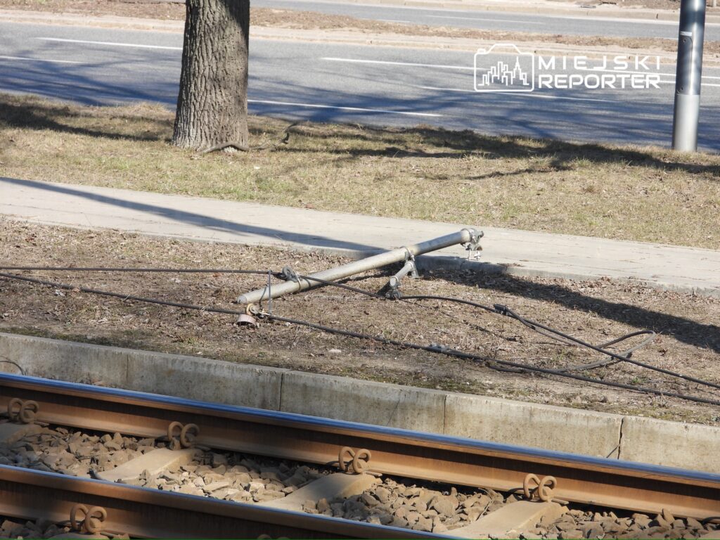 Złamany słup trakcyjny leży na ziemi obok torów tramwajowych, w otoczeniu trawnika i asfaltowej drogi.