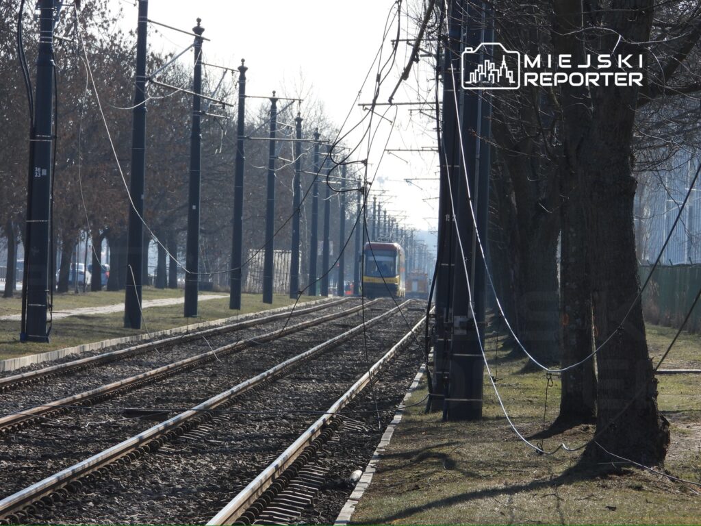 Wzdłuż torów tramwajowych stoją słupy z przewodami, a w tle widoczny jest tramwaj na stacji.