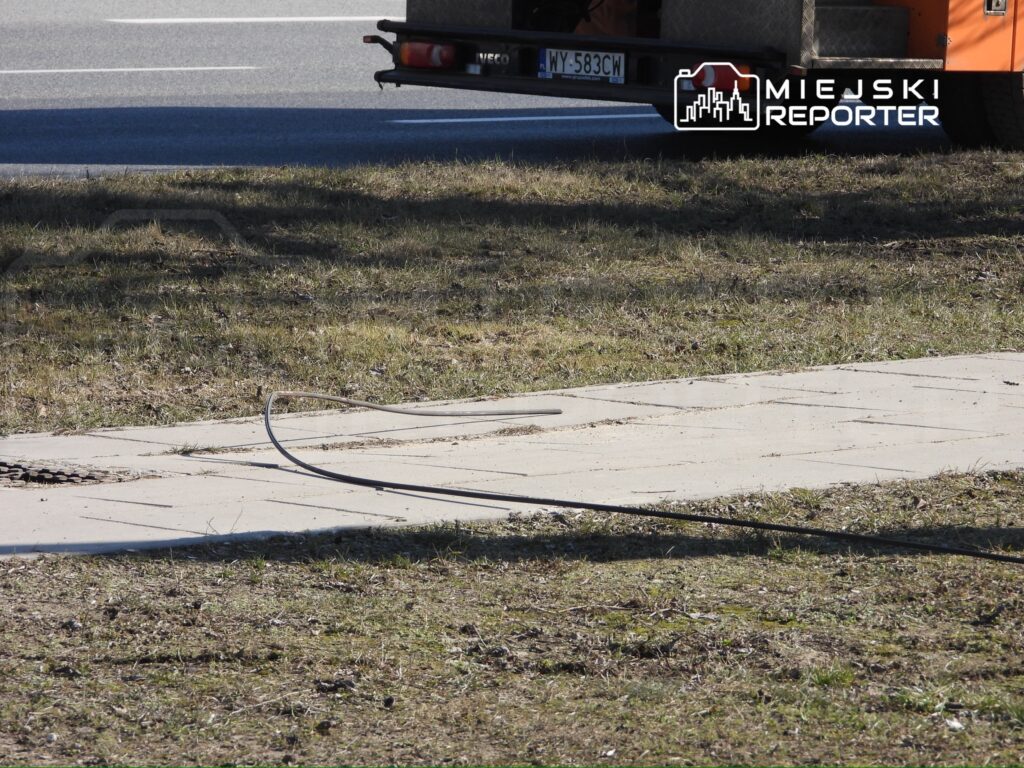 Na trawie przy chodniku leży zwinięty kabel, w tle widoczna droga i pojazd dostawczy.