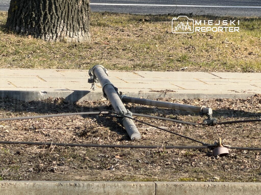 Złamany słup telefoniczny leży na ziemi obok chodnika, otoczony wyschniętą trawą i kablami.