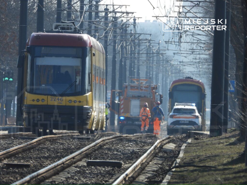 Tramwaj na torach, obok pracownicy w odzieży roboczej oraz pojazdy służbowe w tle.