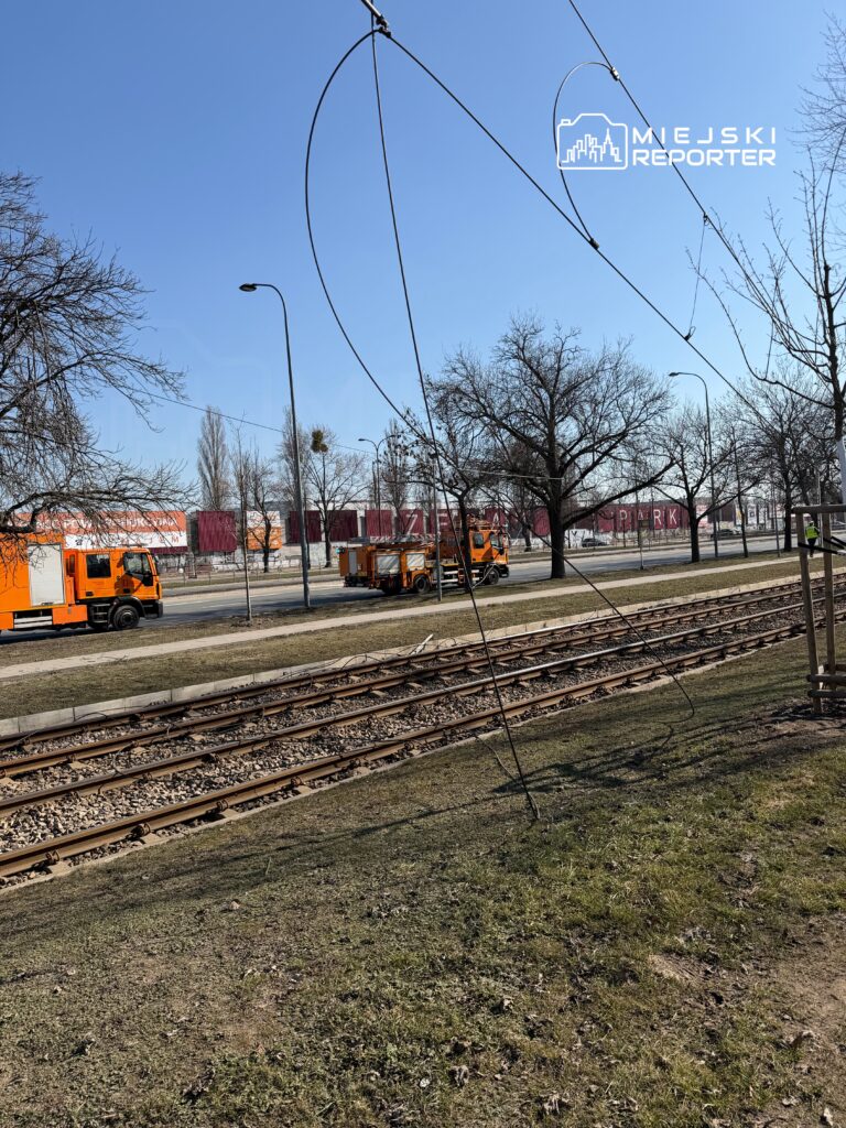 Dwa pomarańczowe pojazdy pracują przy torach tramwajowych w słoneczny dzień, otoczone nagimi drzewami.