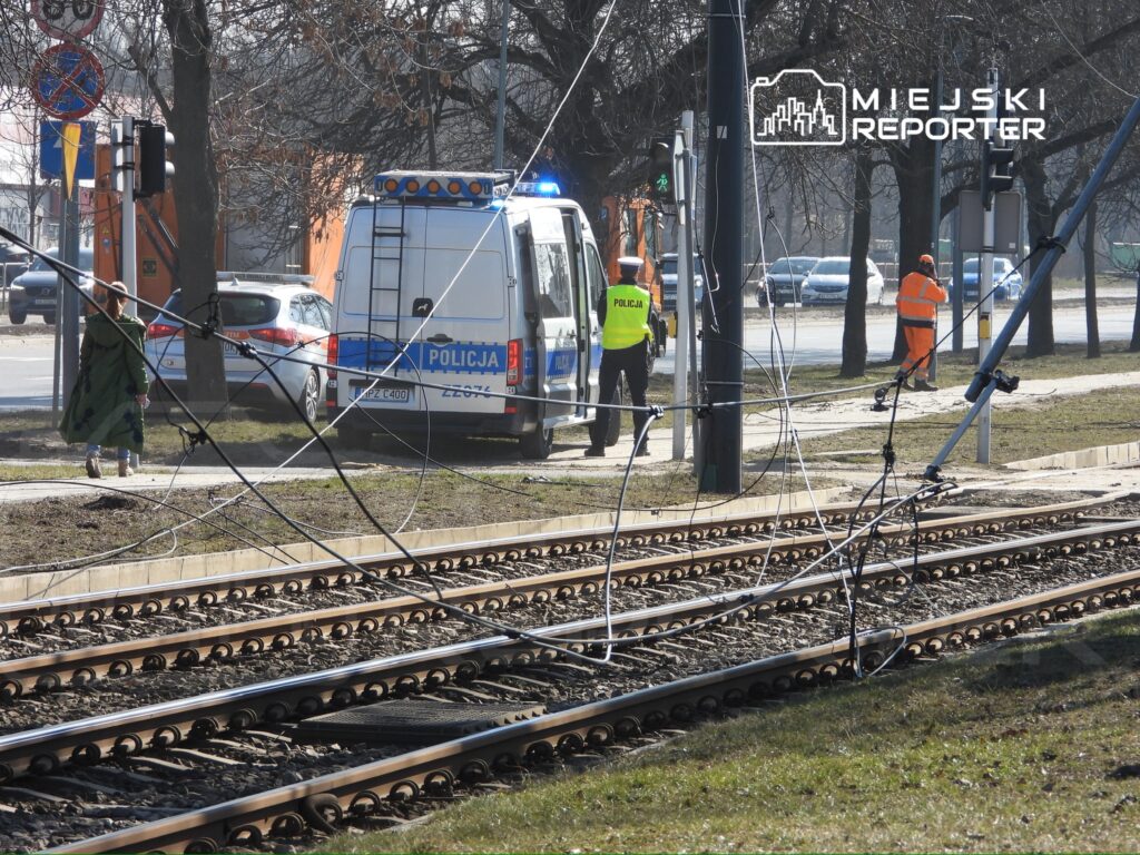 Policjant w kamizelce odblaskowej rozmawia z pracownikami w pomarańczowych ubraniach obok uszkodzonej sieci trakcyjnej.