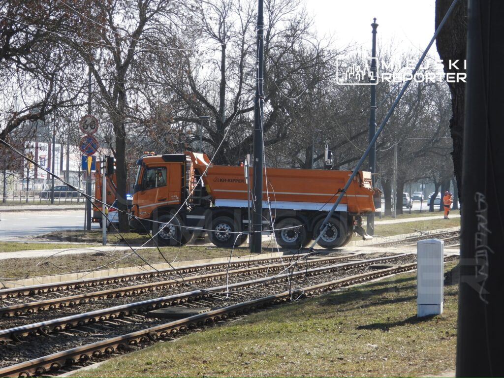 Pomarańczowy samochód ciężarowy z wywrotką stoi obok torów tramwajowych, w otoczeniu drzew i znaków drogowych.