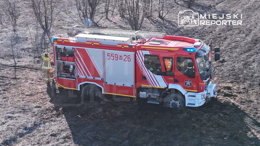Strażak w mundurze z napisem „STRAŻ” obsługuje sprzęt przy wozie strażackim na zniszczonym, poparzonym terenie.