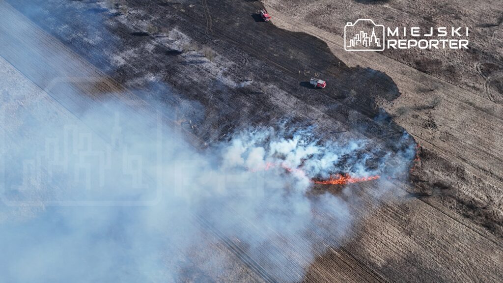 Pożar traw wydobywający się z ziemi na rozległym, poprzecinanym terenie, z widocznymi służbami ratunkowymi w tle.