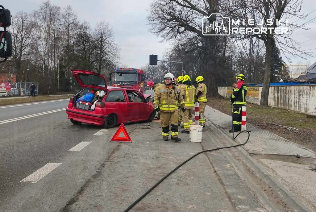 Czerwony samochód osobowy z otwartą klapą bagażnika stoi na poboczu, obok grupy strażaków w specjalistycznych strojach.