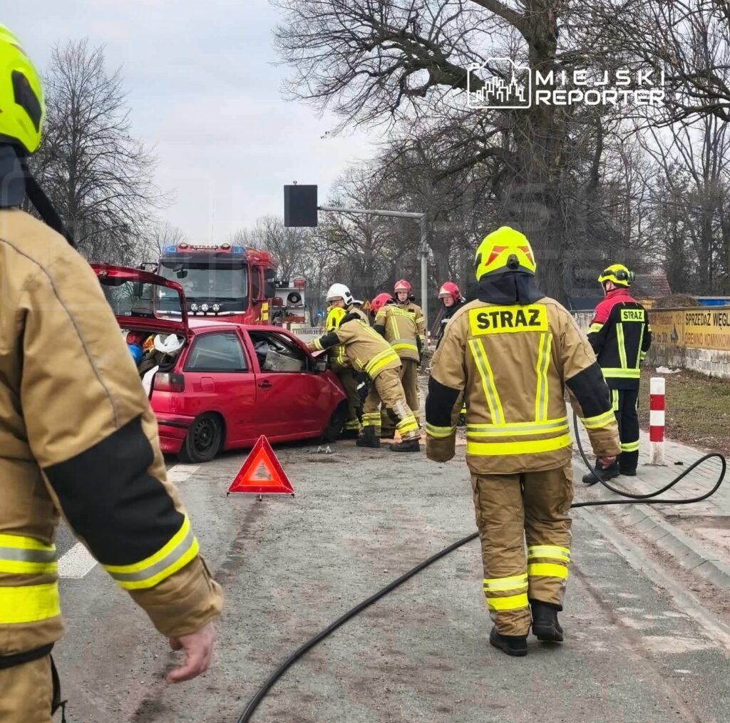Grupa strażaków w żółtych uniformach udziela pomocy poszkodowanemu w czerwonym samochodzie na drodze, obok wozu strażackiego.