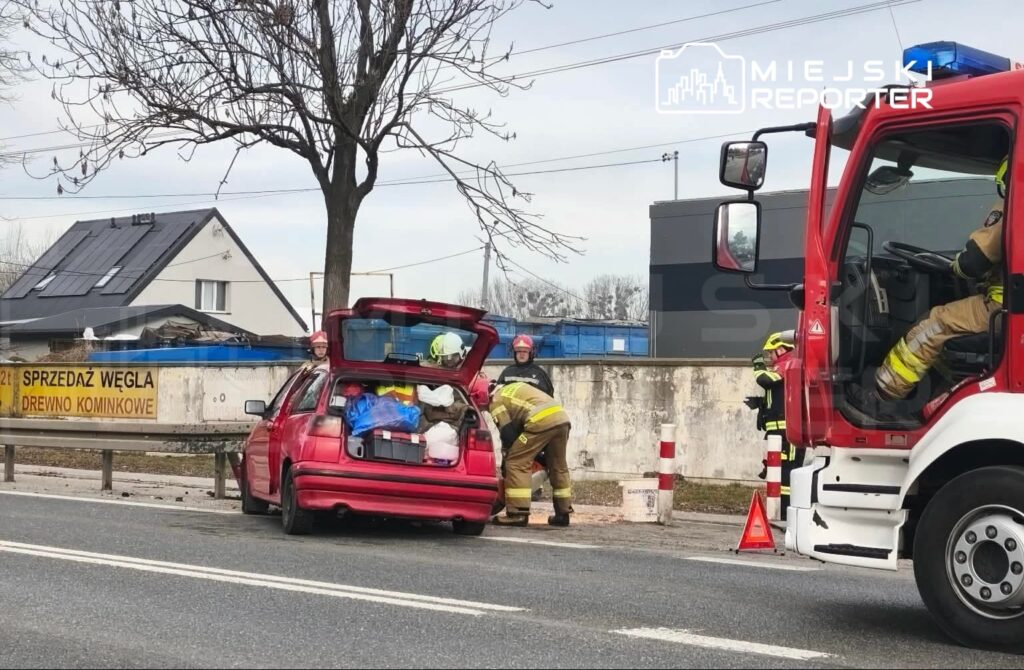 Strażacy przeszukują bagażnik czerwonego samochodu osobowego przy drodze, obok zaparkowanego wozu strażackiego.